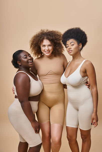 happy multiethnic plus size girlfriends in lingerie posing on beige backdrop, body positive beauty - Foto, Imagem