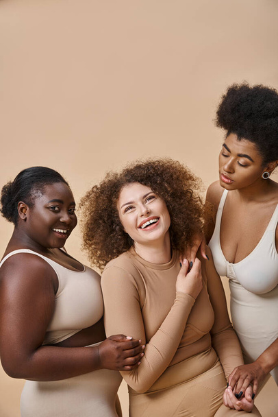 joyful multiethnic plus size girlfriends in lingerie posing on beige backdrop, body positivity - Fotoğraf, Görsel