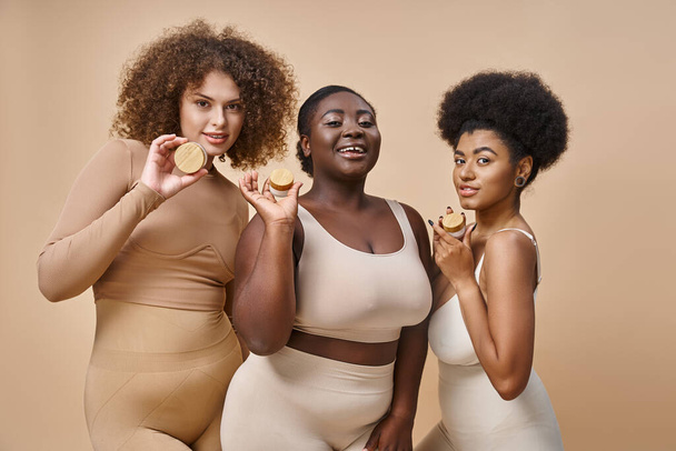 felici donne multietniche plus size in lingerie in posa con barattoli di crema cosmetica su beige, skincare - Foto, immagini