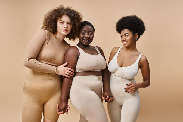 sonriente más tamaño multirracial mujeres en lencería posando en beige, belleza y positividad corporal - Foto, imagen