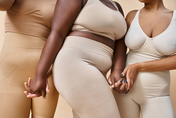 cropped view of plus size and curvy women in underwear holding hands on beige, body positivity - Foto, Imagen