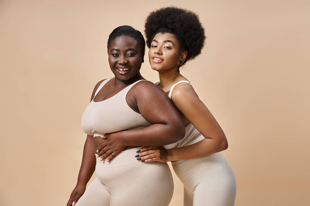 encantador más tamaño afroamericano mujeres en ropa interior mirando a la cámara en beige, positividad del cuerpo - Foto, imagen