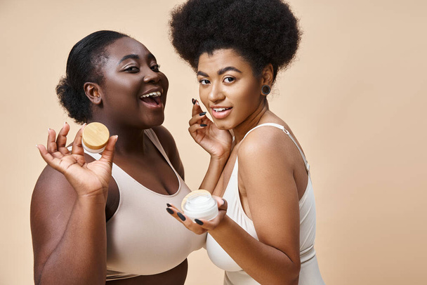 radosny afrykański plus rozmiar kobiety w bieliźnie pozowanie z kosmetycznym kremem na beżowy, pielęgnacja skóry - Zdjęcie, obraz