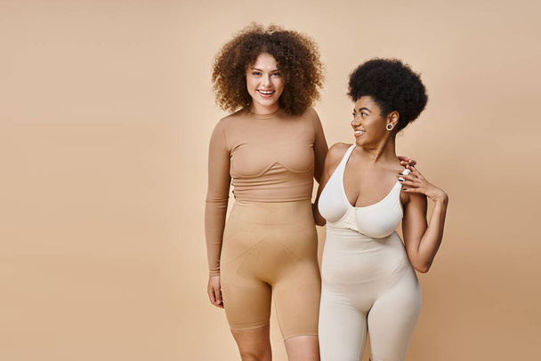 usmívající se multikulturní tělo pozitivní ženy ve spodním prádle stojící na béžové, přírodní plus velikost krásy - Fotografie, Obrázek