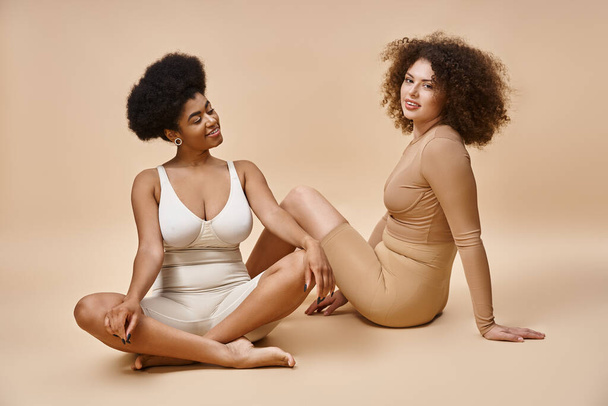 cheerful multicultural plus size girlfriends in lingerie sitting on beige backdrop, natural beauty - Foto, immagini