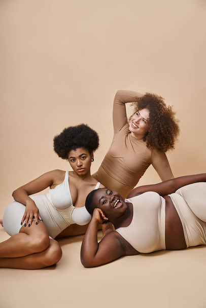 happy multiracial body positive women in lingerie posing on beige backdrop, natural beauty - 写真・画像