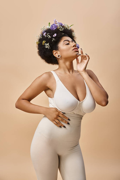 sensuele Afrikaanse Amerikaanse vrouw met kleurrijke bloemen in haar poseren in lingerie op beige achtergrond - Foto, afbeelding