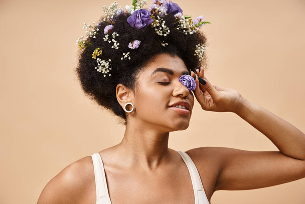 ευτυχισμένη Αφροαμερικανή γυναίκα με floral διακόσμηση σε μαλλιά και λουλούδια κοντά στο πρόσωπο σε μπεζ, φυσική ομορφιά - Φωτογραφία, εικόνα