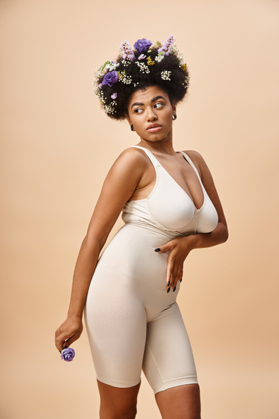 curvy femme afro-américaine avec des fleurs dans les cheveux posant en lingerie sur beige, beauté naturelle - Photo, image