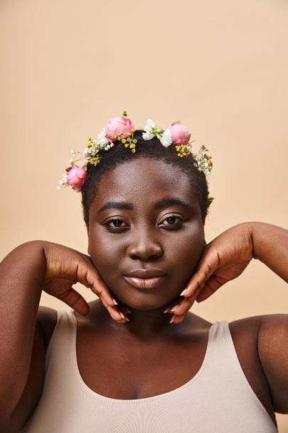 charming plus size african american woman with flowers in hair and hands near face on beige - Fotó, kép