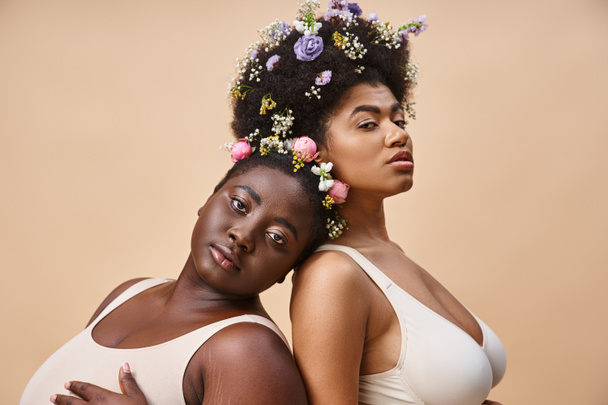 charmant Afrikaans-Amerikaanse vrouwen met bloemen in haar poseren in ondergoed op beige, plus size schoonheid - Foto, afbeelding