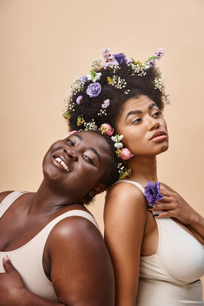 charmante afrikanisch-amerikanische Frauen mit Blumen im Haar posieren in Unterwäsche auf beige, plus size beauty - Foto, Bild