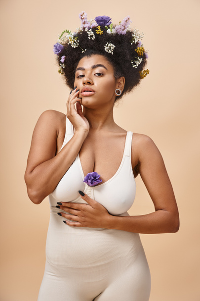 İç çamaşırlı, bej renkli, doğal güzellikteki saçlarında çiçeklerle poz veren Afro-Amerikalı kadın. - Fotoğraf, Görsel