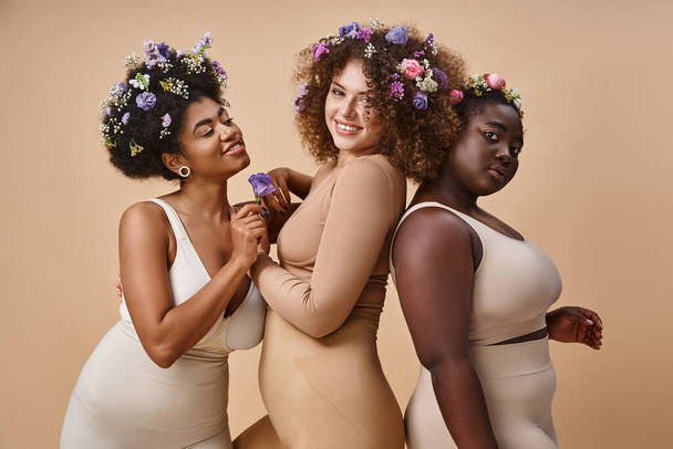 feliz multirracial plus size mulheres em lingerie com flores coloridas no cabelo em bege, charme curvilíneo - Foto, Imagem
