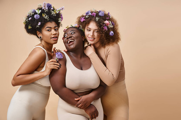 happy multiracial plus size women in lingerie with colorful flowers in hair on beige, curvy charm - Photo, Image