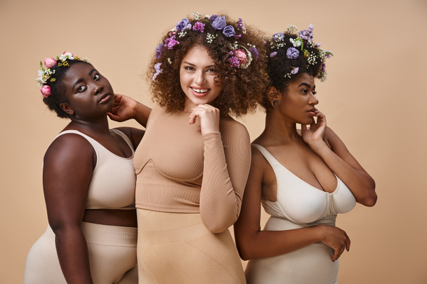 joyful multiethnic girlfriends in lingerie with colorful flowers in hair on beige, plus size beauty - Photo, Image