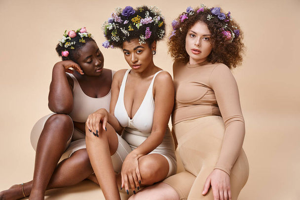 cuerpo positivo mujeres multiétnicas en lencería con flores en el pelo sentado en beige, belleza natural - Foto, imagen