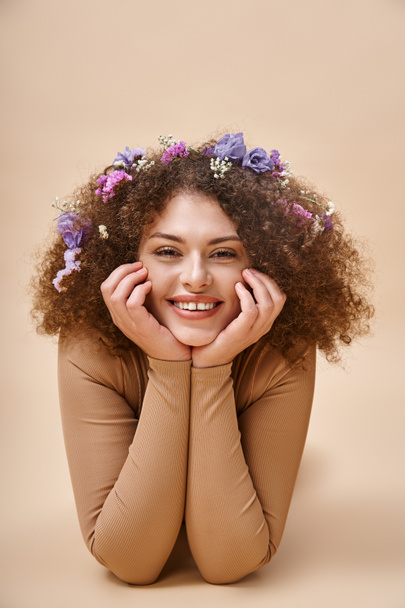 Porträt einer hübschen und fröhlichen Frau mit bunten Blumen im welligen Haar auf beige, natürliche Schönheit - Foto, Bild