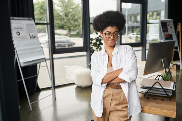 με αυτοπεποίθηση χαμογελαστή Αφροαμερικανίδα επιχειρηματίας με γυαλιά ηλίου που στέκεται με διπλωμένα χέρια στο γραφείο - Φωτογραφία, εικόνα
