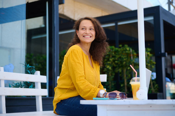 Autentický portrét multietnicky usmívající se šťastné krásné ženy, která se dívá stranou, jak sedí na letní terase kavárny. Příjemná podnikatelka pracující venku na notebooku. Lidi. Životní styl - Fotografie, Obrázek