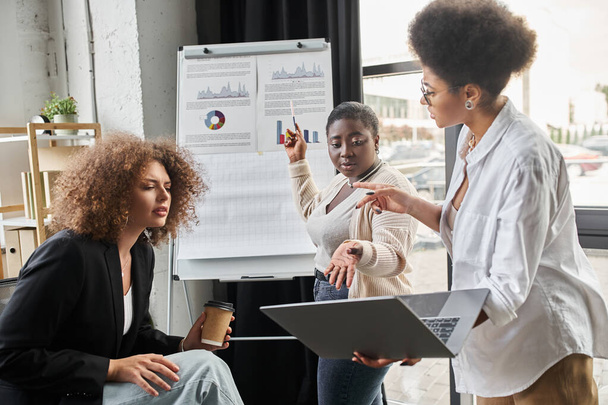 equipe criativa de mulheres de negócios multiétnicas falando perto de laptop e flip chart no escritório, inicialização - Foto, Imagem