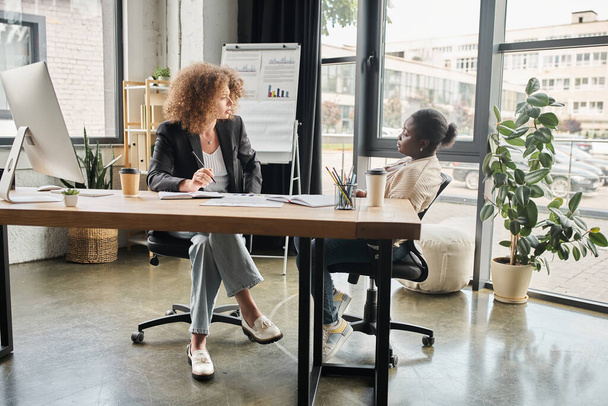 femme d'affaires interracial parler près de moniteur d'ordinateur et de documents sur le lieu de travail dans le bureau moderne - Photo, image
