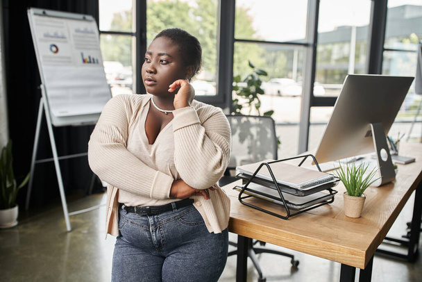 przemyślany afrykański amerykański plus rozmiar businesswoman odwracając wzrok podczas stojąc w nowoczesny biuro - Zdjęcie, obraz