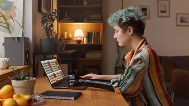 Widok z boku talia w górę strzał kaukaski freelance dziewczyna z protezą ramię za pomocą laptopa podczas siedzenia przy biurku przez okno w przytulnym biurze - Materiał filmowy, wideo