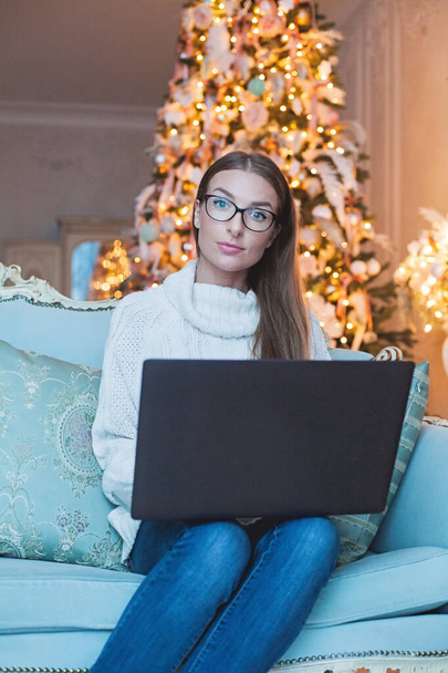 Aranyos hölgy fehér pulóver segítségével laptop ül a kék kanapén előtt a karácsonyfa otthon - Fotó, kép