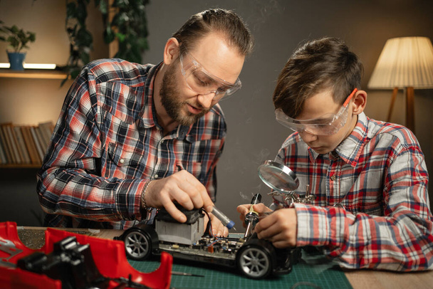 Vader die zijn zoon les geeft voor het solderen en repareren van op afstand bestuurde auto 's thuis. Man en jongen maken stuk speelgoed. Kopieerruimte - Foto, afbeelding