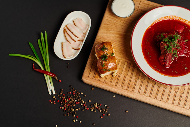свіжий український буряковий суп біля часникових булочок, свинячого салату та зеленої цибулі на обробній дошці на чорному - Фото, зображення