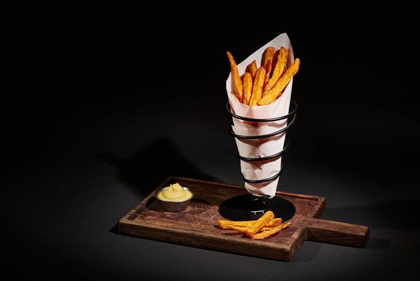 batatas fritas crocantes gourmet dentro do cone de papel perto do molho de imersão na placa de corte de madeira - Foto, Imagem