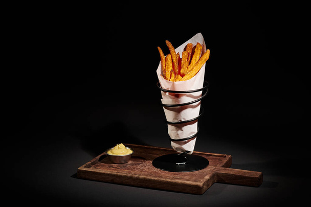 удобная еда, хрустящая картошка фри внутри бумажного конуса рядом с соусом для окунания на деревянной доске - Фото, изображение