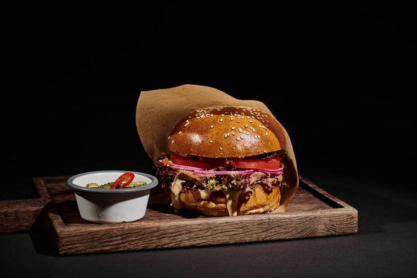 delizioso cheeseburger con panino al sesamo, manzo e sottaceti come contorno su vassoio di legno su nero - Foto, immagini