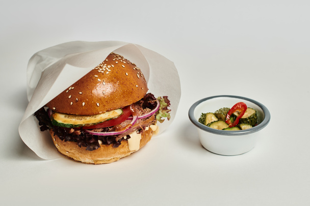вкусный гамбургер с булочкой кунжута, говядиной и огурцами в качестве гарнира на деревянном подносе на сером фоне - Фото, изображение