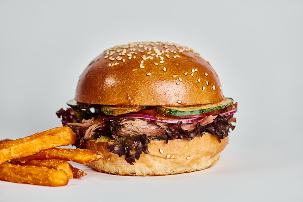 hambúrguer saboroso com carne, cebola vermelha, tomate e pão de gergelim perto de batatas fritas em pano de fundo cinza - Foto, Imagem