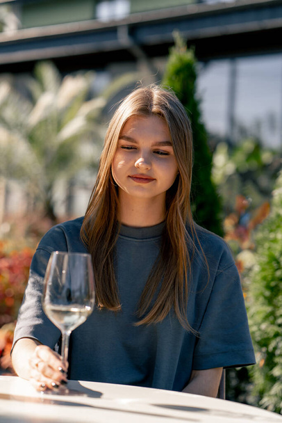 Ein junges schönes Mädchen hält ein Glas Wein in der Hand und untersucht es sorgfältig, um die Qualität des Getränks zu bestimmen - Foto, Bild