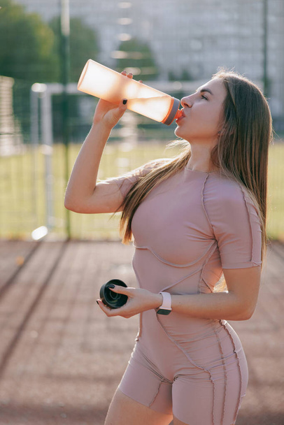 Jonge vrouw drinkt water uit de fles na de training op het sportveld. Vrouw oefent buiten. Lichamelijke training en gezonde levensstijl. - Foto, afbeelding