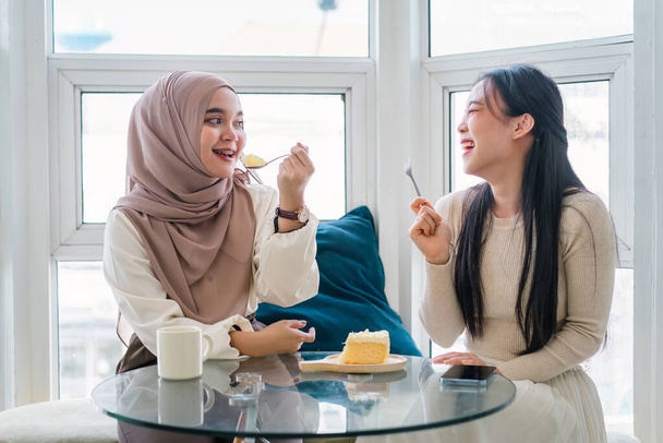 Dos amigos multiétnicos disfrutando de pastel y café juntos en una cafetería, charlando y riendo - Foto, Imagen