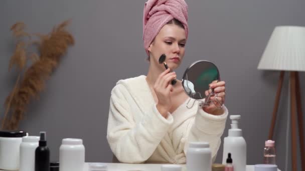 Jonge vrouw met een perfecte huid draagt witte badjas en handdoek op het hoofd maken van gezichtsmassage met behulp van een jade gezicht roller voor spiegel in haar woonkamer. Kopieerruimte - Video