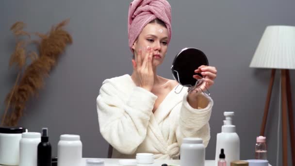 Junge hübsche Frau mit Bademantel und Handtuch im Haar, die Feuchtigkeitscreme auf ihr Gesicht aufträgt. Rituale zur Hautpflege. Kopierraum - Filmmaterial, Video
