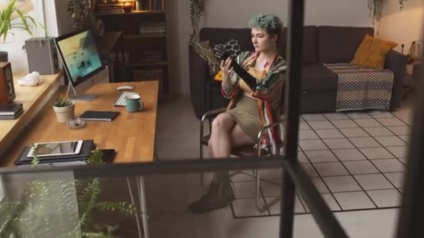 Hidas valkoihoinen tyttö vammainen istuu tuolilla puinen työpöytä ikkunalta viihtyisässä asunnossa katsoen huolellisesti hänen proteesi käsivarteen - Materiaali, video