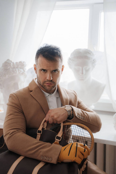 Богатый человек в старом денежном стиле держит сумку и теннисную ракетку в мастерской скульпторов - Фото, изображение