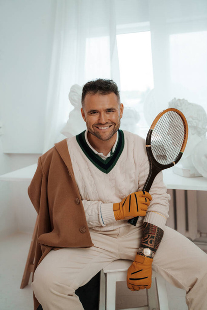 Очаровательный мужчина в старом денежном стиле, с очаровательной улыбкой, позирует с теннисной ракеткой среди древнегреческих скульптур - Фото, изображение