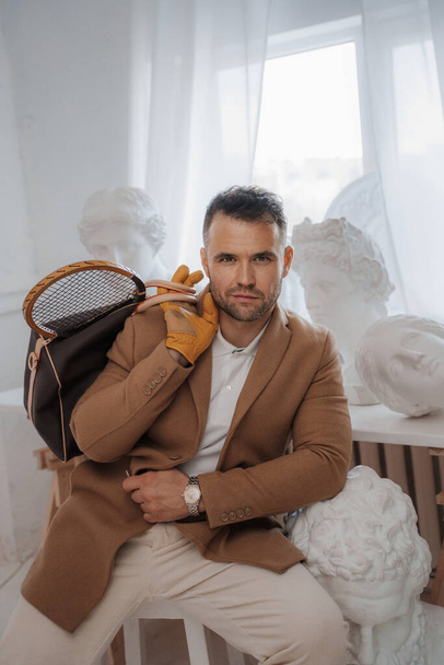 Stylish male model embodies the old-money look, posing with a bag and tennis racket among classical Greek sculptures in a sculptors atelier - Photo, Image