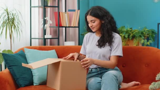 Ευτυχισμένη Ινδή νεαρή γυναίκα ξεπακετάρει δέμα παράδοσης κάθεται στο σπίτι. Χαμογελώντας ικανοποιημένοι Arabian Hindu κορίτσι shopper online shop πελάτη άνοιγμα χαρτονένιο κουτί λάβετε δώρο αγοράς με γρήγορη ταχυδρομική αποστολή - Πλάνα, βίντεο