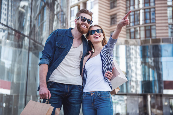 Beautiful young couple in sun glasses and with shopping bags holding hands, looking away and smiling while walking down the street - Photo, Image