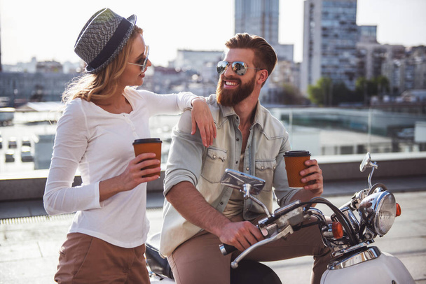 Beautiful young couple in sun glasses is drinking coffee, talking and smiling while sitting on a scooter outdoors - Photo, Image
