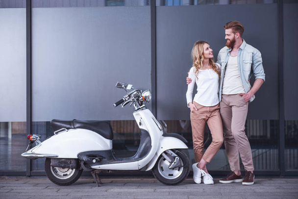 Красивая молодая пара обнимается, смотрит друг на друга и улыбается, стоя возле скутера на улице
 - Фото, изображение