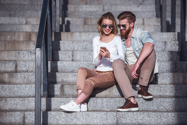Beautiful young couple in sun glasses is using a smart phone and smiling while sitting on stairs outdoors - Photo, Image